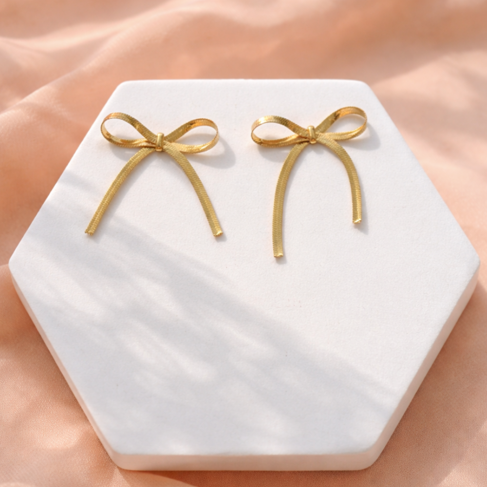 Gold bow-shaped earrings on a hexagonal white stone surface with a soft pink background