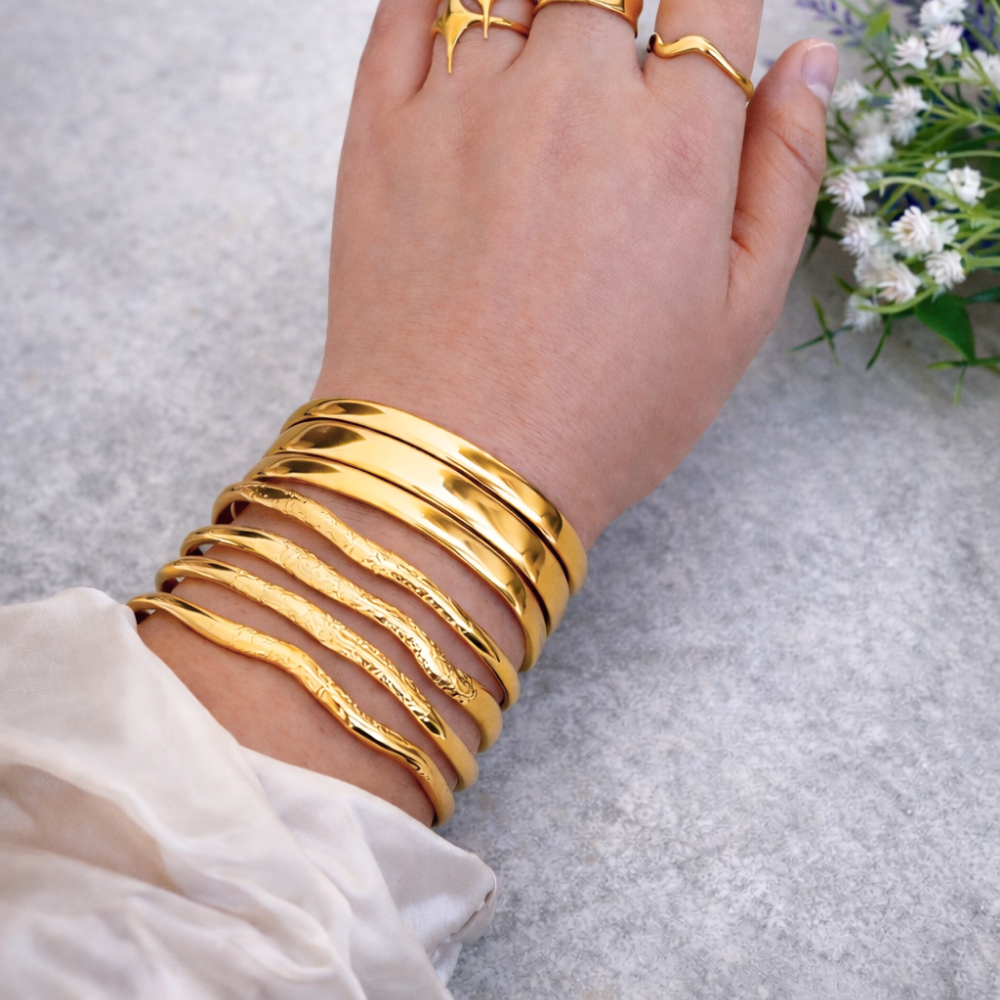Hand wearing multiple gold bracelets on a light gray background with a small bouquet of flowers.