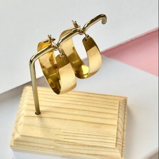 A pair of gold plated hoop earrings displayed on a wooden stand against a white and pink background.
