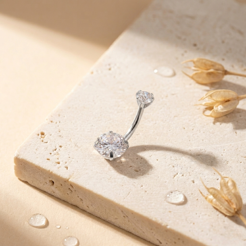 Silver belly button ring on a beige stone surface with dried flowers.