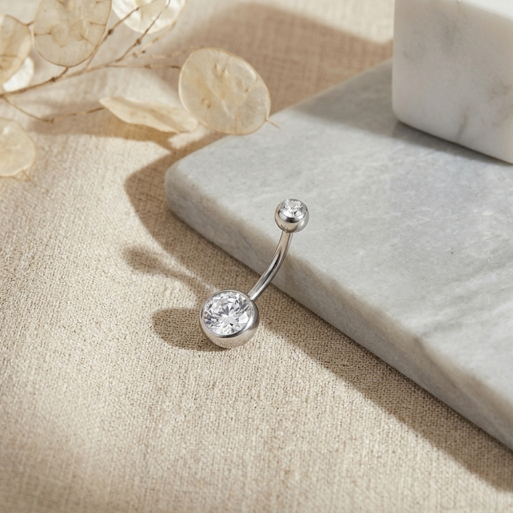 Silver belly button ring with clear gemstone on a beige fabric background