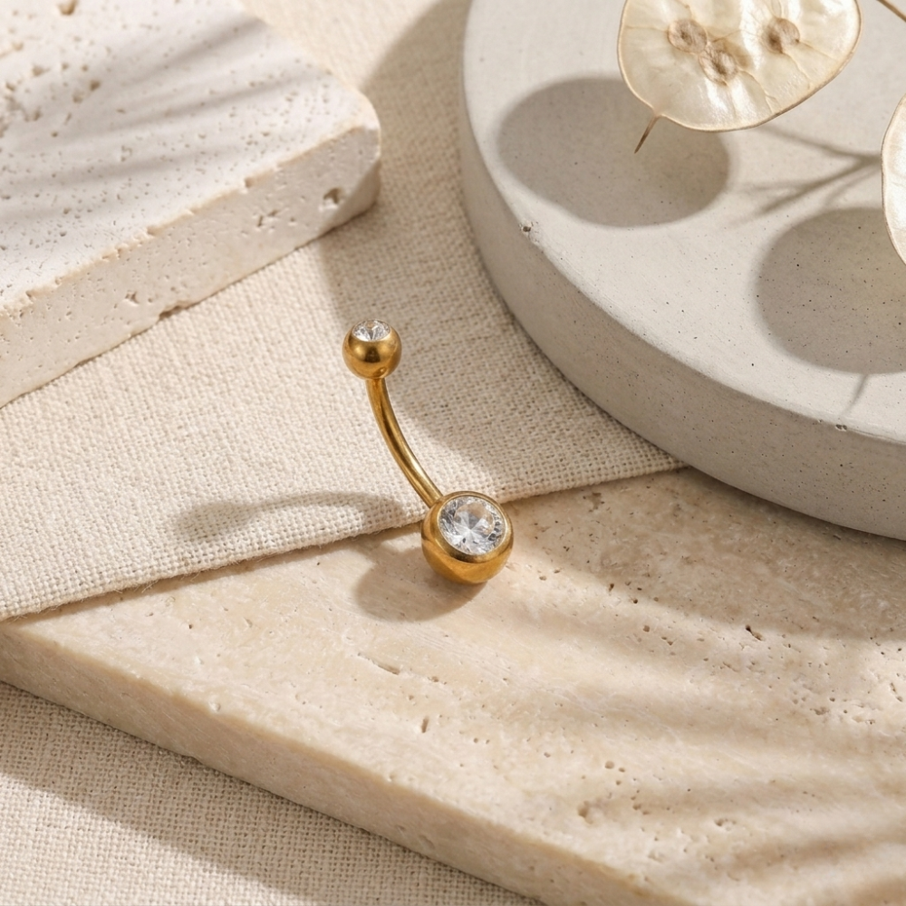Gold belly button ring with clear gemstone on a beige surface